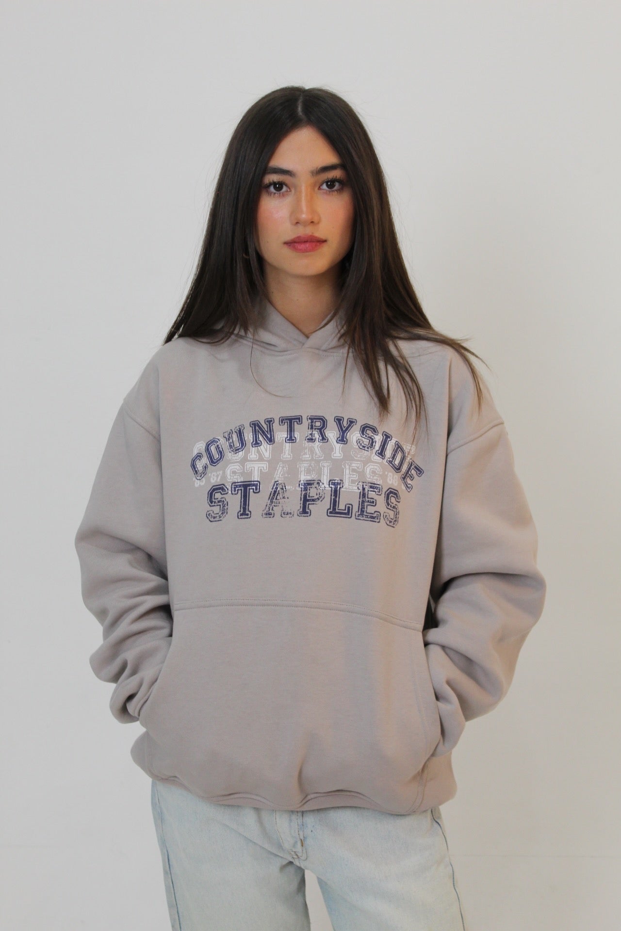 Model wearing gray hoodie with hood up, Countryside Staples arched logo across chest.